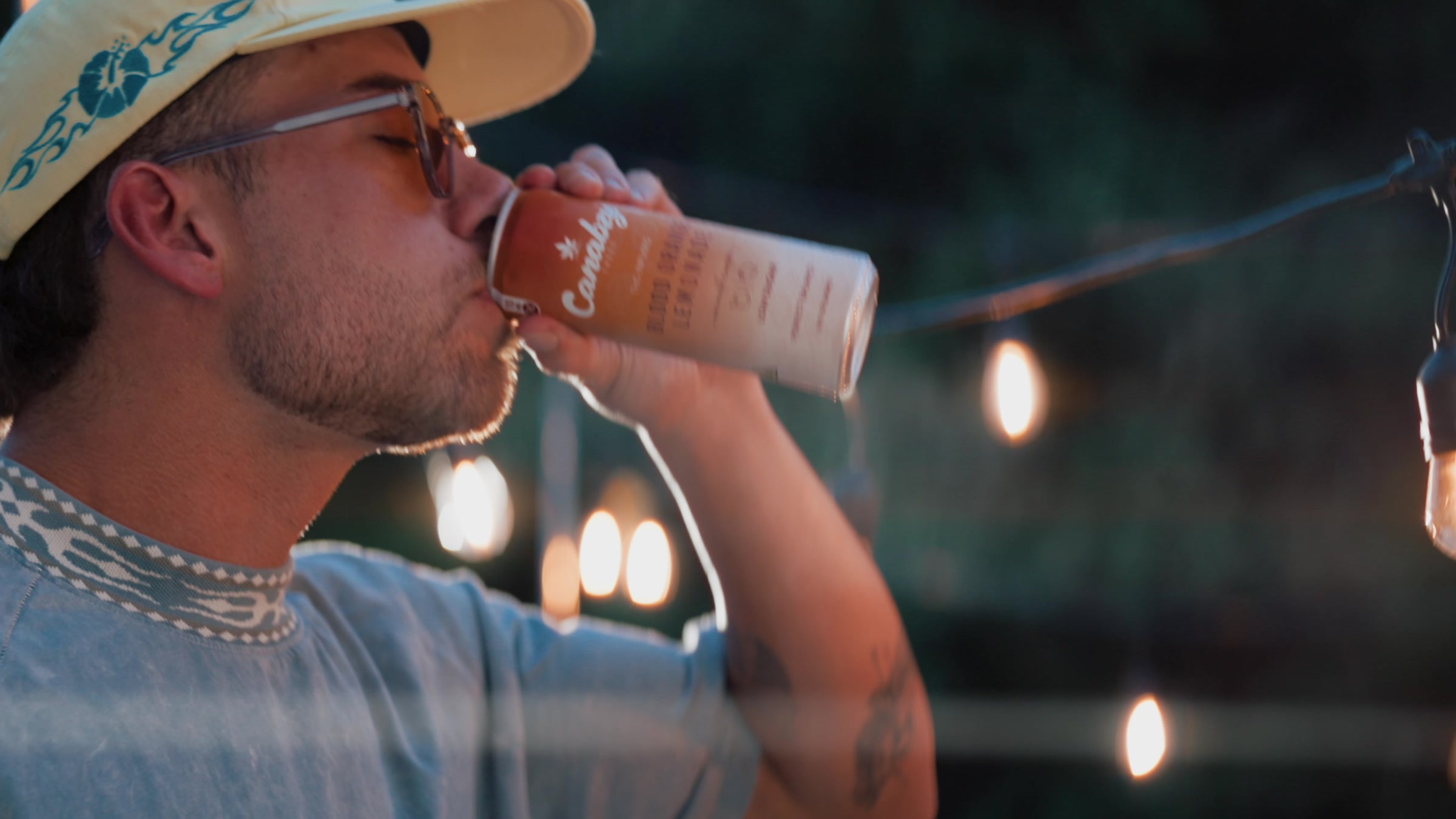 A hand holding a Canabzy Blood Orange Lemonade can beside a lit outdoor grill at dusk